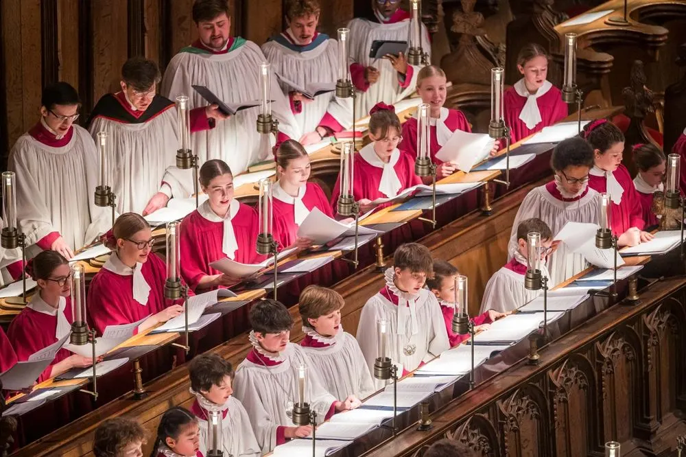 Choir members in red and white robes singing from music stands in a cathedral choir stalls.