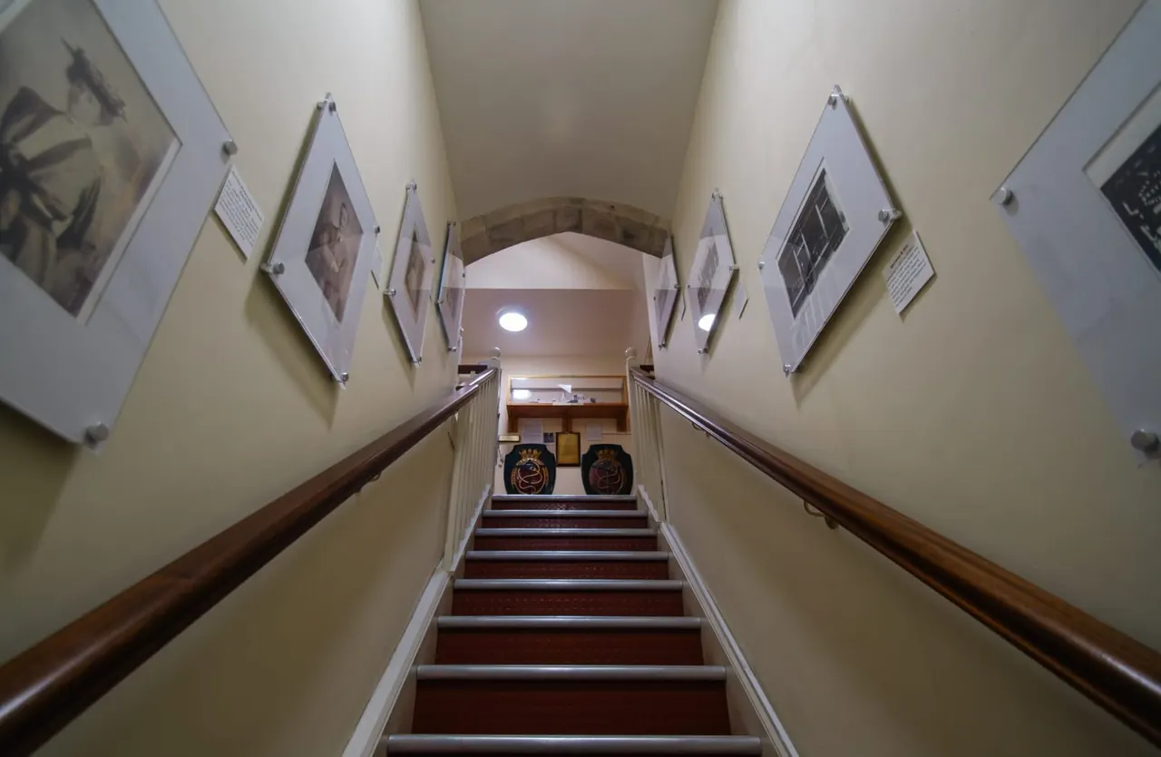 Staircase with mounted photographs and information panels on either side, leading to a landing with decorative items on display.