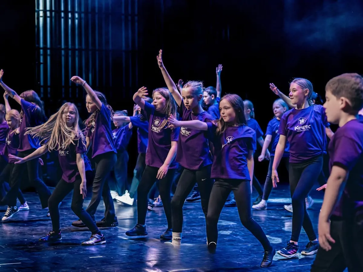 Group of young dancers performing choreographed routine on stage in purple-lit theatre with blue lighting.