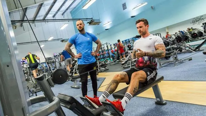 People exercising in a busy gym with weights, cardio machines and training equipment visible throughout the facility.