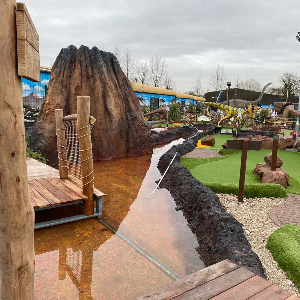 Giant dinosaur-themed miniature golf course with a large tree stump structure, themed obstacles, and tropical landscaping beneath overcast skies.