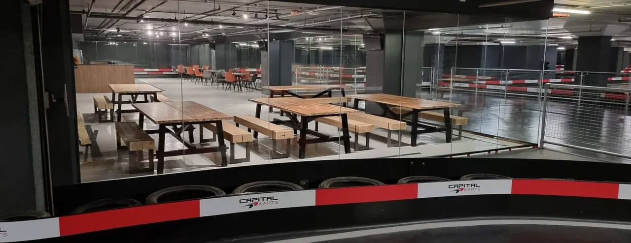 Modern indoor kart racing facility with wooden picnic tables and benches in a glass-walled social area.