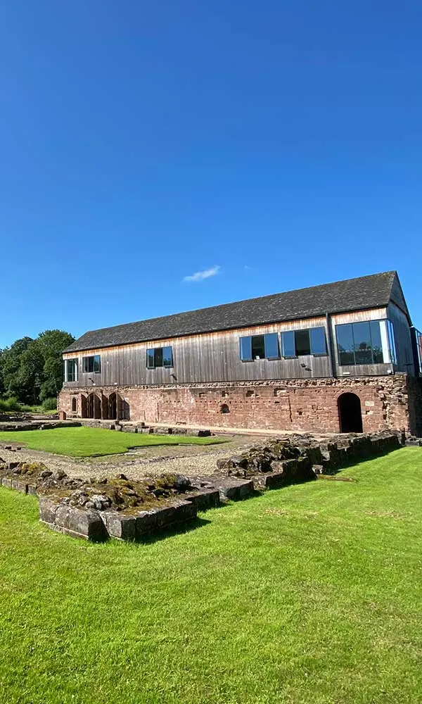 Modern building with wooden cladding built on historic red sandstone ruins, set in manicured green lawns under blue sky.