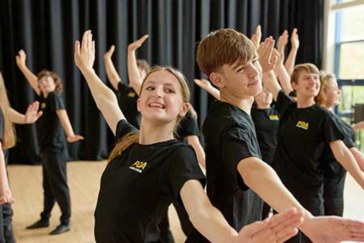 Group of young performers in black t-shirts dancing with raised arms on a stage with dark curtains behind them.