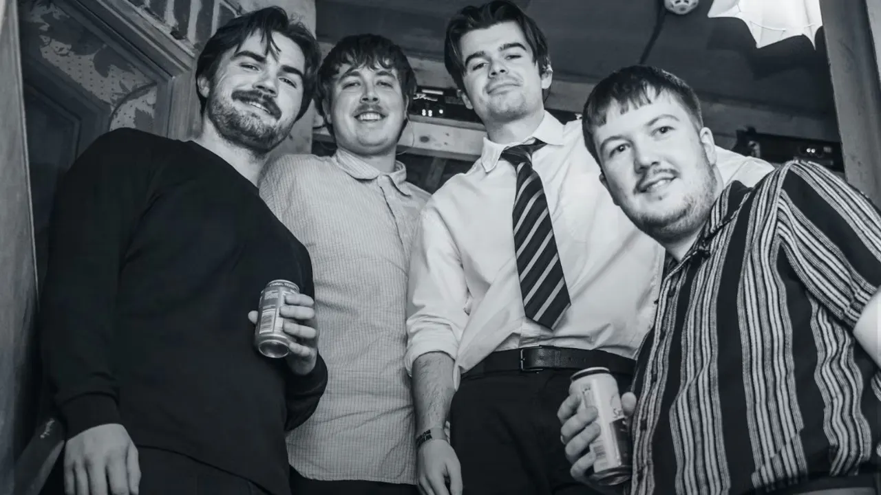 Four men smiling at the camera indoors, holding drinks at what appears to be a pub or bar.