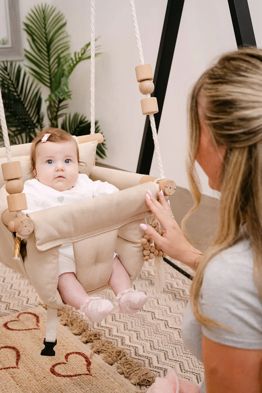 Adult woman interacting with a baby in a suspended fabric swing with wooden beads and straps.