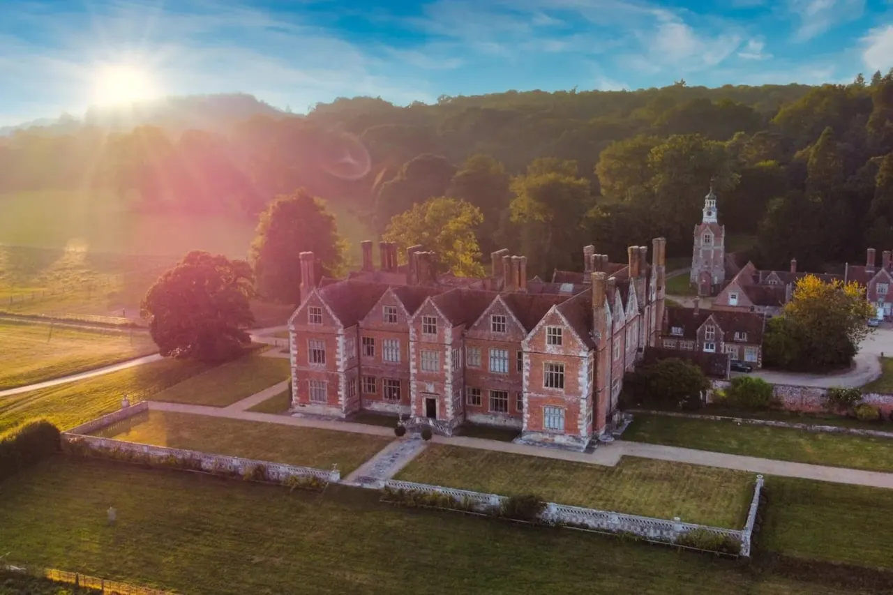 Aerial view of a red-brick Tudor mansion with manicured gardens, surrounded by woodland and rolling countryside at sunrise.