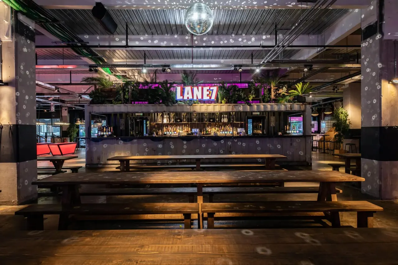 Modern bowling alley bar and lounge with wooden seating, illuminated signage, and disco ball overhead lighting.