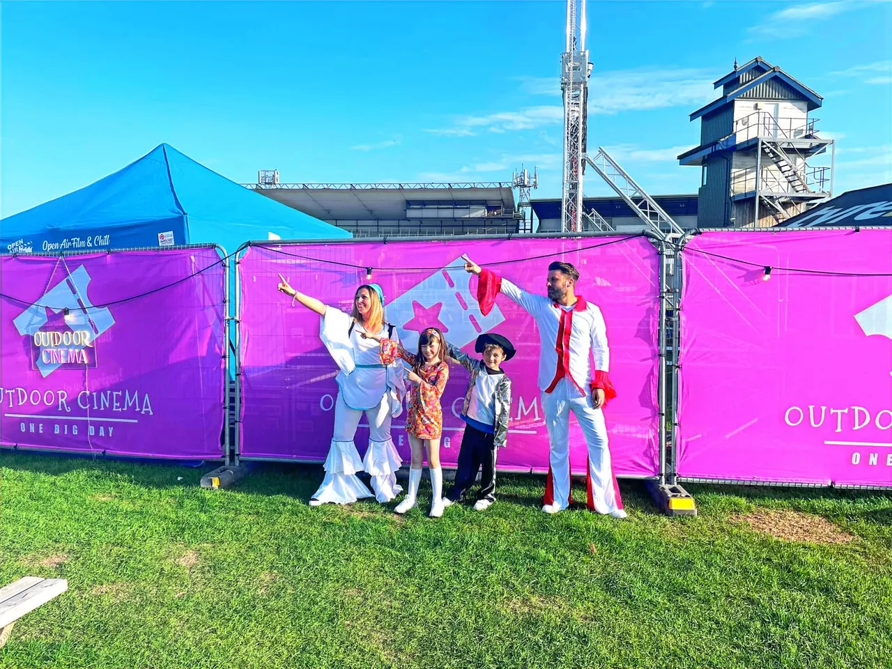 A family in festive fancy dress posing outside a bright pink outdoor cinema tent at a sports ground venue.