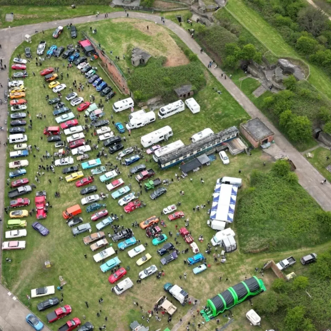 Aerial view of a car and vehicle rally on grass beside a historic fort, with hundreds of colourful vehicles and caravans displayed.