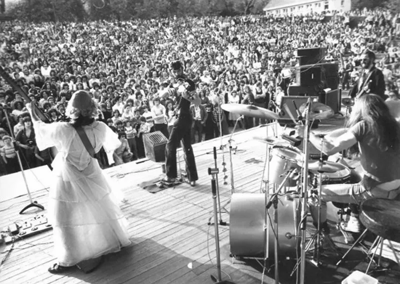 Band performing on stage to a large outdoor crowd at a park venue.