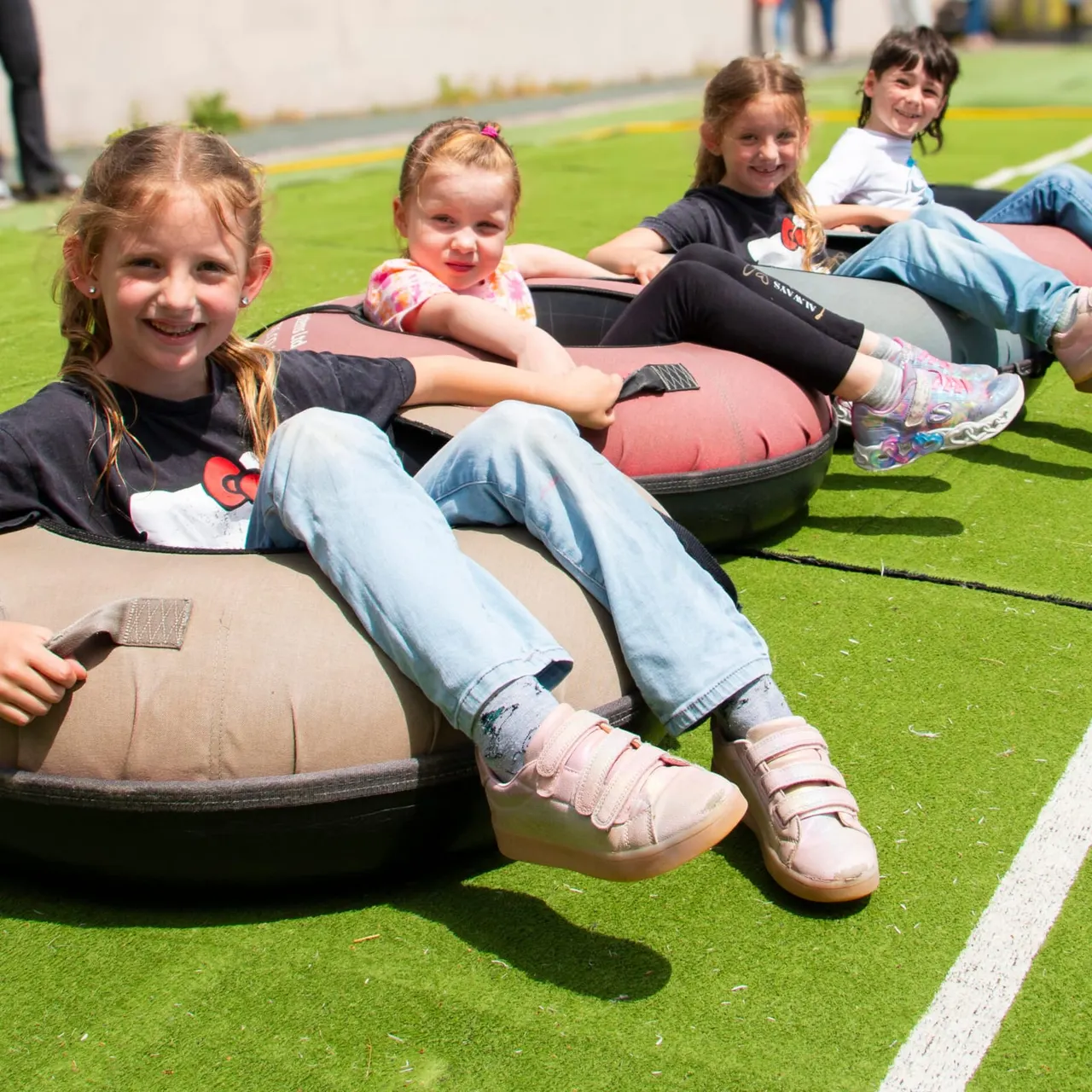 Four children sitting in tyre tubes on artificial grass, smiling at the camera during a leisure activity.