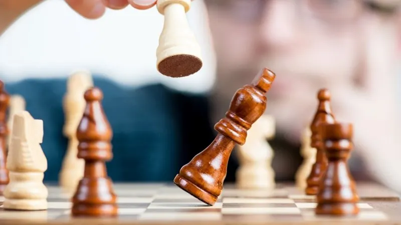A hand holds a wooden chess piece above a board mid-game with other pieces visible.