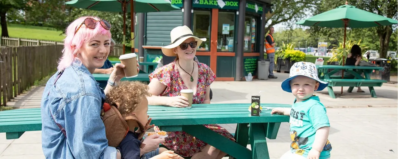 Two adults and two young children enjoy drinks and food at a green picnic table in a sunny park, with a café building and umbrellas visible behind them.