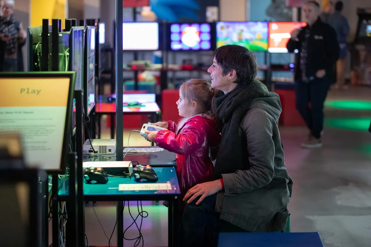 Adult and child playing an interactive video game console together in a busy arcade hall with multiple screens and other visitors in the background.