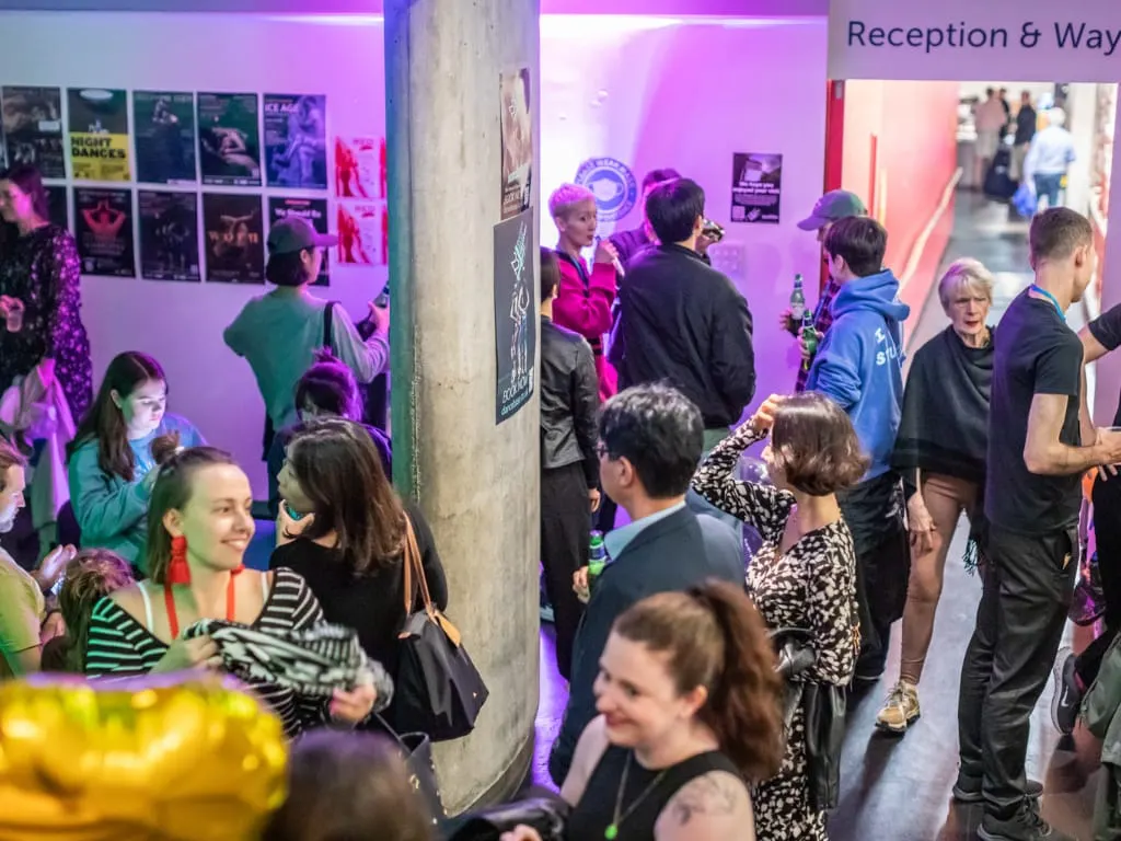 Groups of people socialising and dancing in a vibrant purple and pink lit indoor venue.