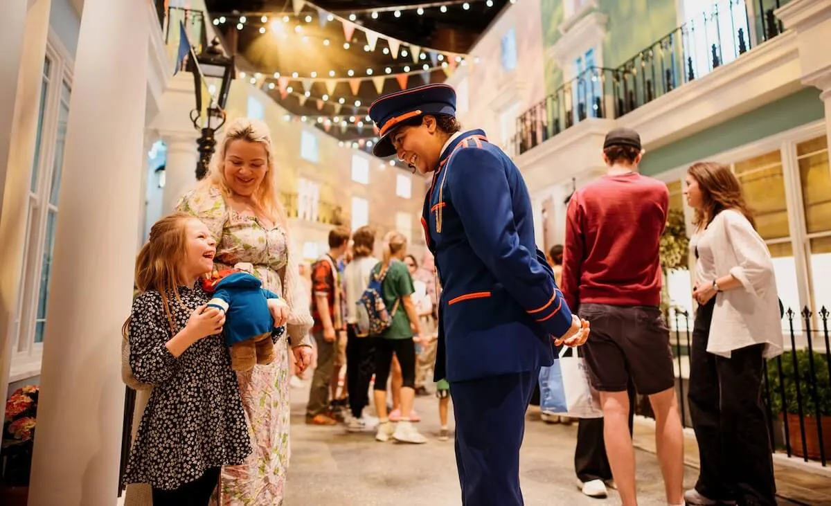 A uniformed staff member interacts with a young girl holding a teddy bear in a decorated courtyard with festive lights and other visitors.