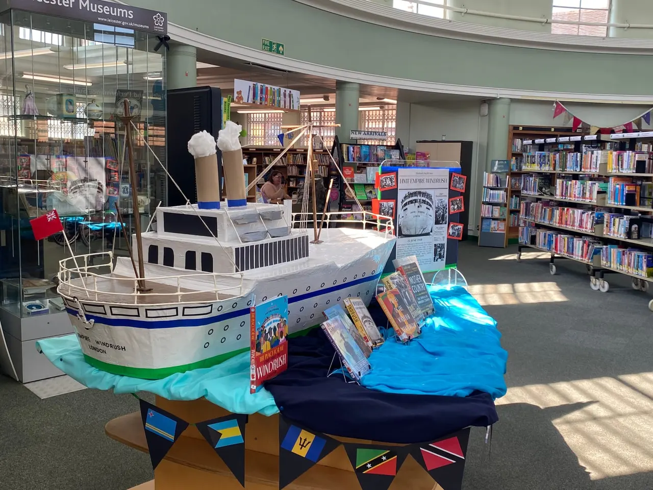 A library interior displays a large model ship exhibition with a wooden sailing vessel model, blue water feature, and maritime-themed decorations including flags and books about the Windrush.