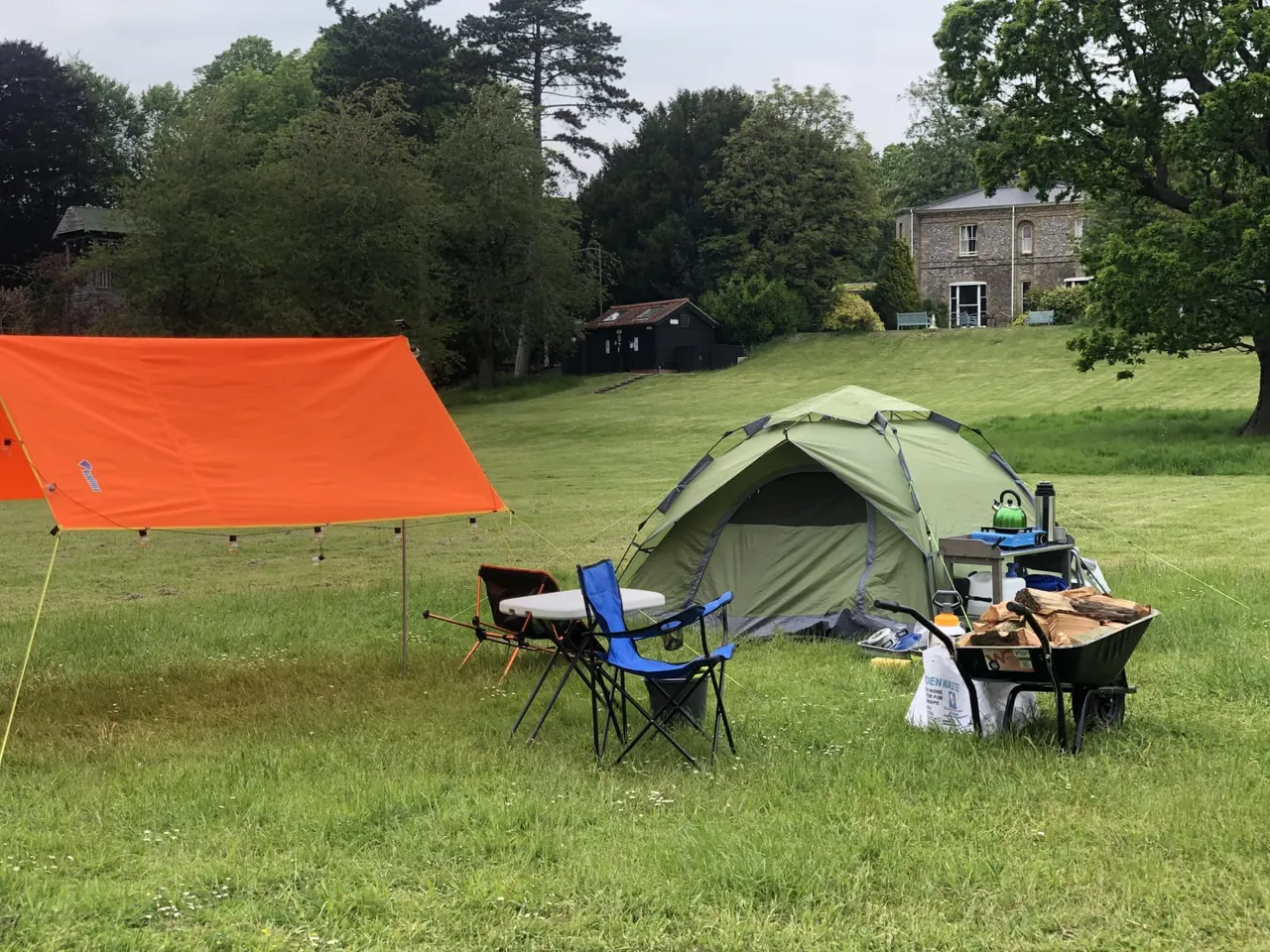 Camping setup on a grassy field with a green tent, orange tarp shelter, camping chairs, and a fire pit, with historic buildings visible behind.