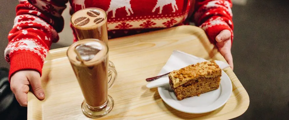 Person in red festive jumper holding wooden tray with hot drinks and slice of cake.