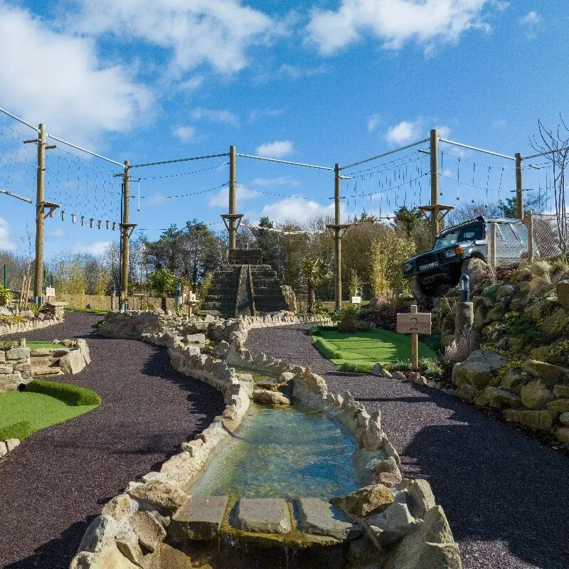 An outdoor adventure course with a small stream, wooden poles with overhead nets, and a vehicle suspended on a ramp against a blue sky.