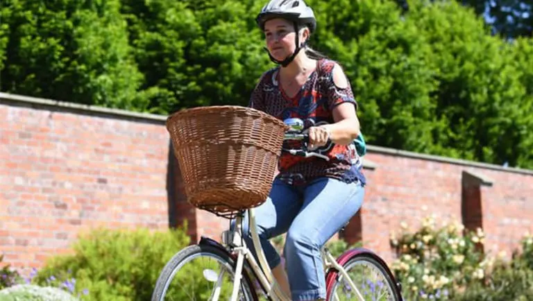 Woman wearing a white helmet riding a bicycle with a wicker basket, past a brick wall and green foliage.
