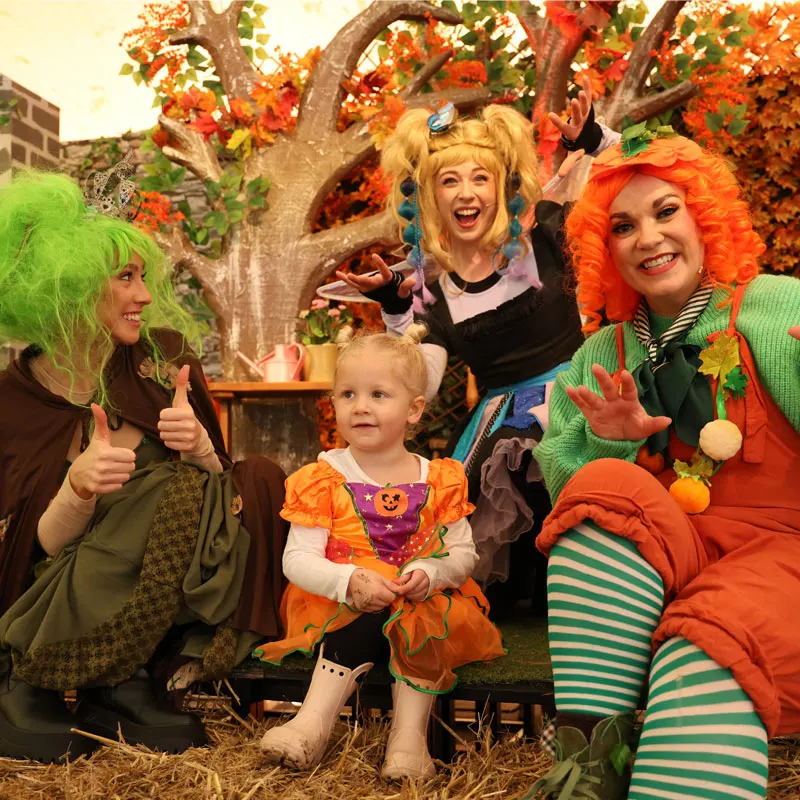 Three adults in colourful wigs and Halloween costumes pose with a toddler in a pumpkin outfit under an autumn tree.