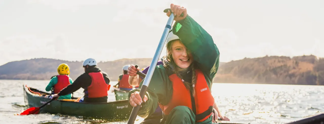 Child in orange life jacket paddling a canoe on a lake with hills in the background, smiling at camera.