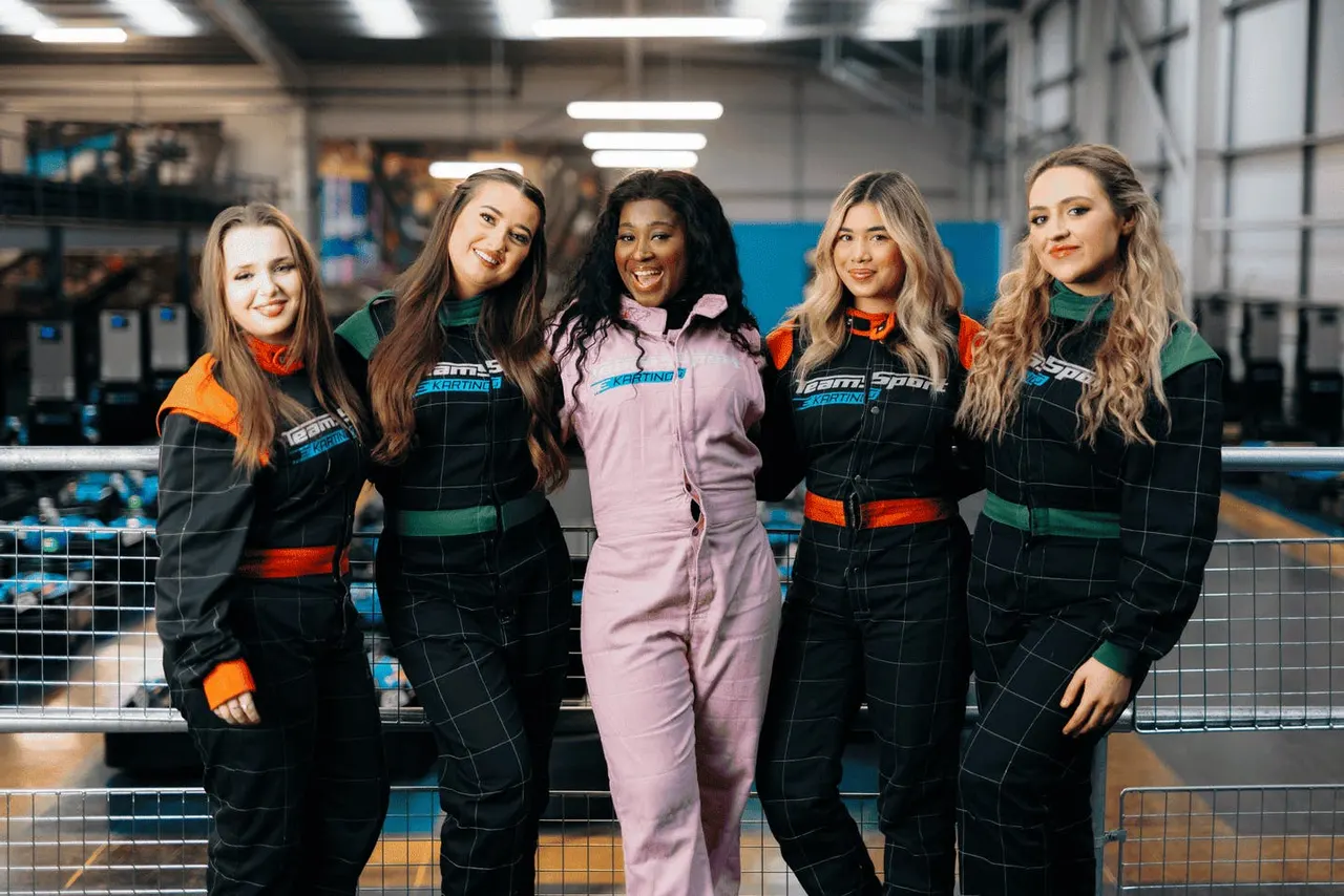 Five young women in racing suits stand together at an indoor go-kart track.