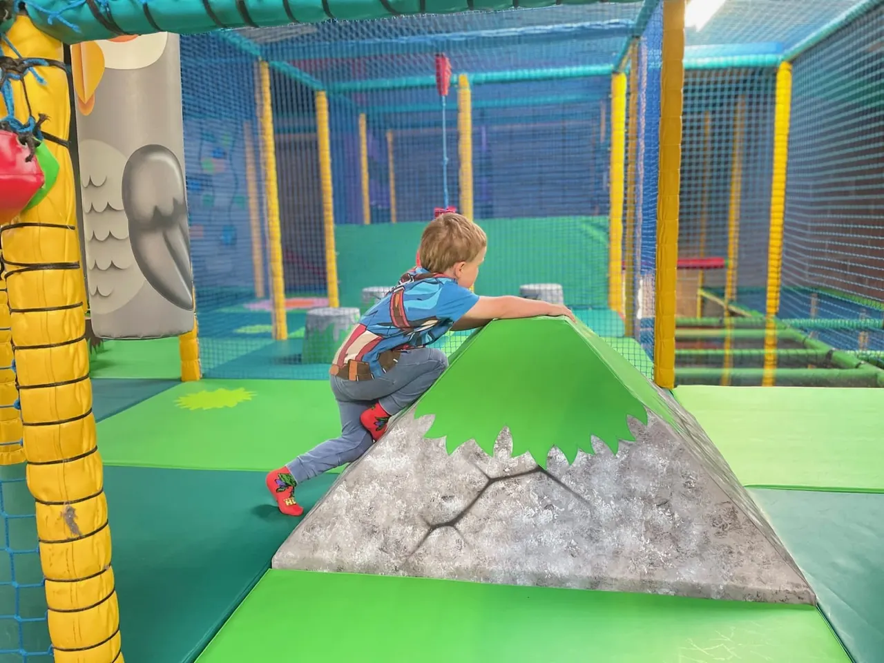 Young child climbing over a rock formation inside an indoor play centre with blue netting and colourful equipment.