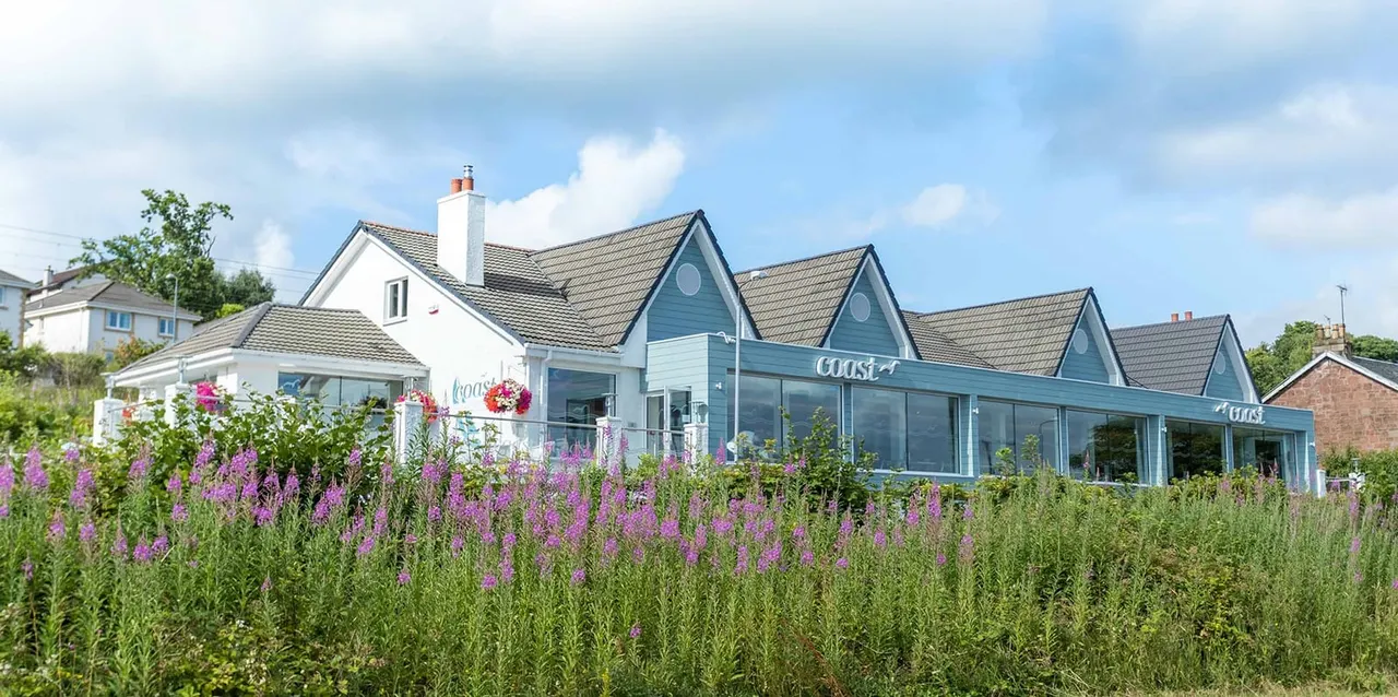 Modern coastal leisure building with blue-grey cladding surrounded by purple flowering plants and hanging baskets on a sunny day.