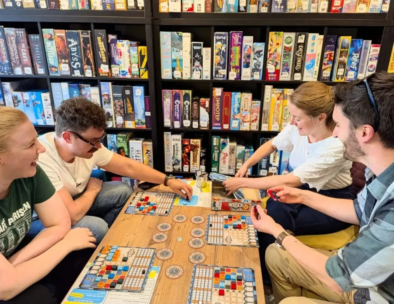 Four people playing board games together at a table with shelves of boxed games displayed behind them.