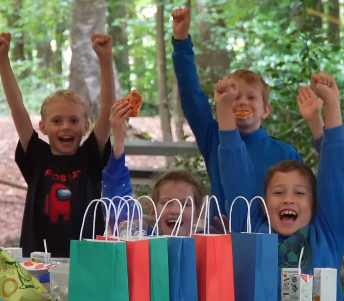 Children at an outdoor woodland picnic table celebrating with arms raised and food, surrounded by colourful party bags.