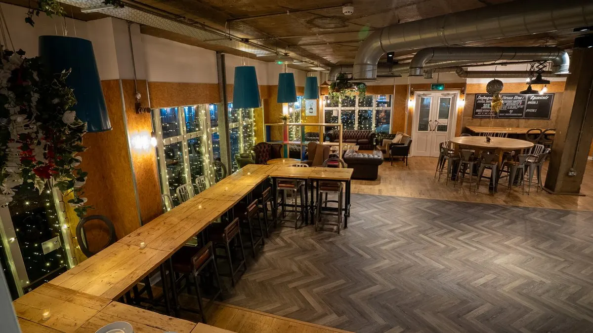 Interior of a modern bar and lounge space with wooden high-top tables, seating areas, and illuminated wine display cabinets.