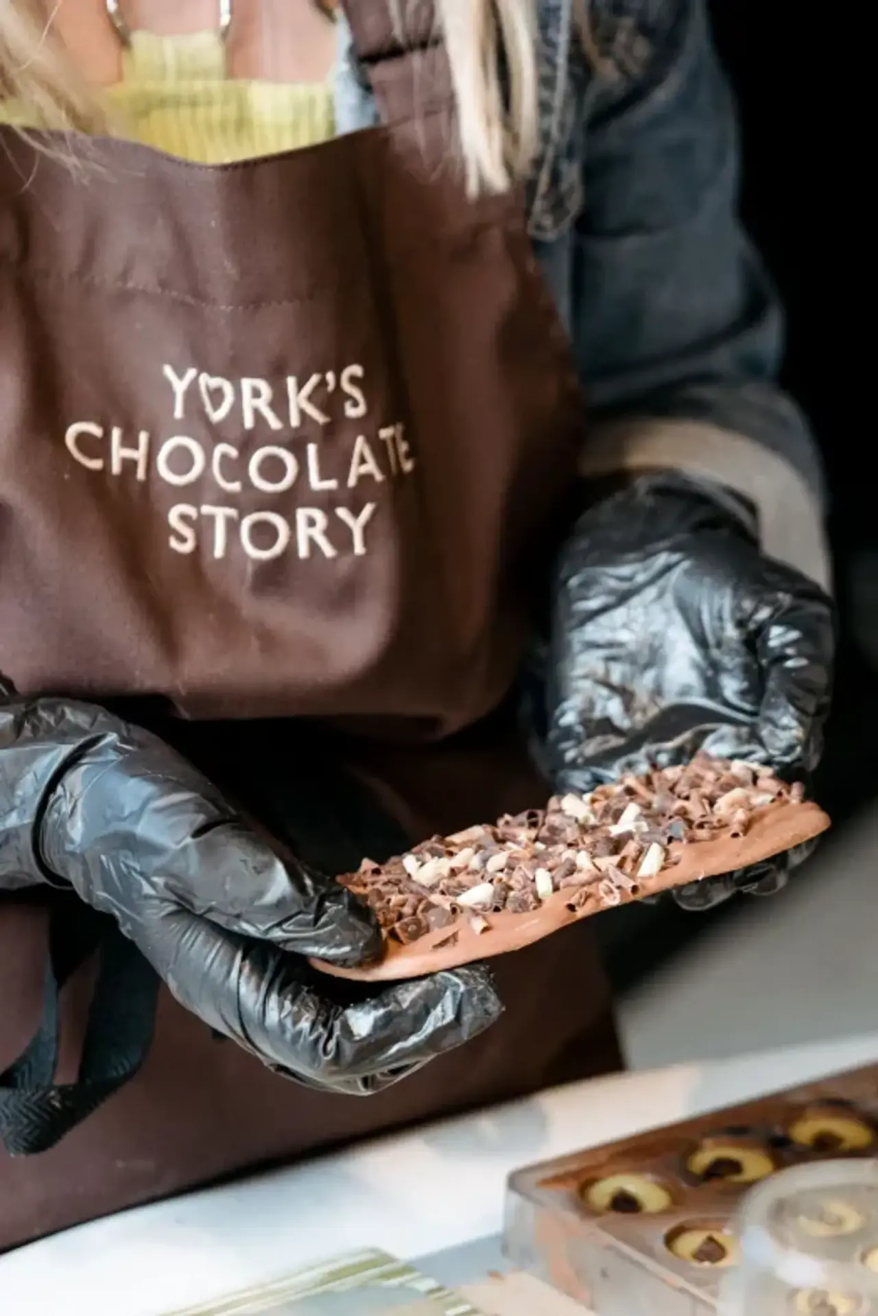 Staff member wearing black gloves holding a chocolate treat topped with nuts and sprinkles at a chocolate attraction.