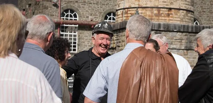 A man in mining hard hat and uniform smiles whilst chatting with visitors outside a stone building with mining equipment.