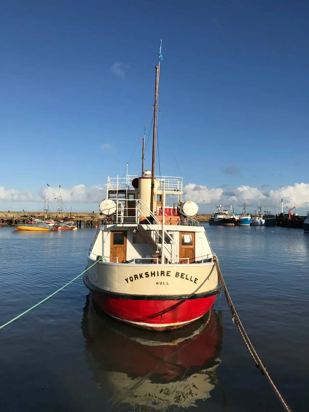 Traditional fishing vessel with red hull and cream superstructure moored in a working harbour under clear blue sky.