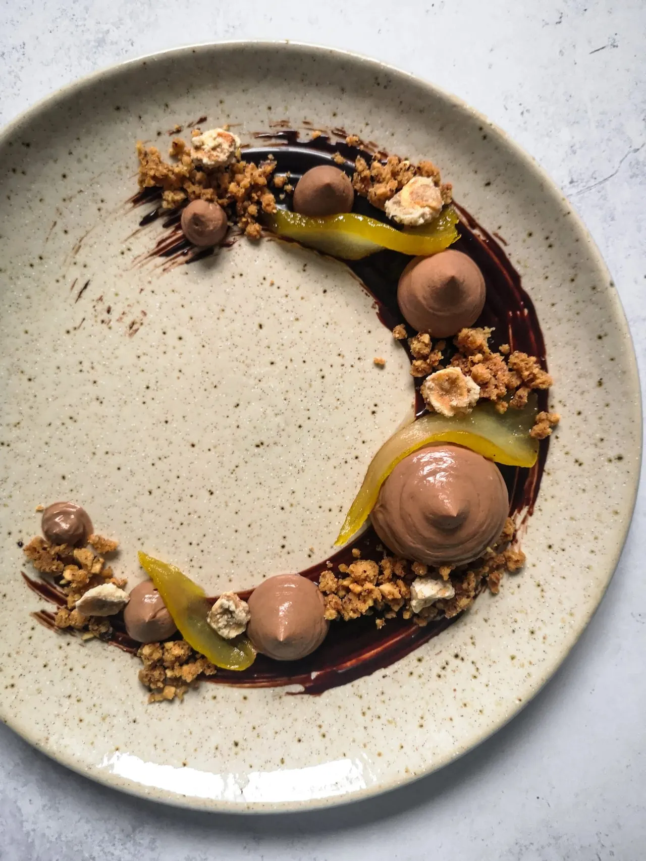 Artfully plated chocolate dessert with chocolate mousse spheres, pistachio elements, crumble, and dark chocolate sauce on a speckled cream plate.