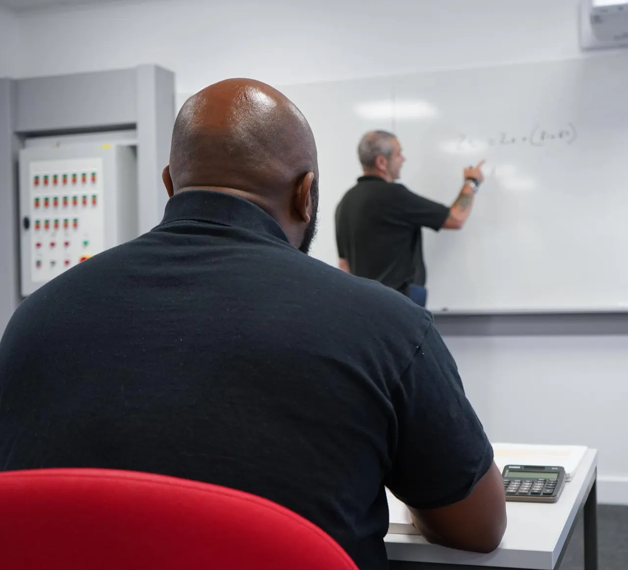 Two men in a training session, one seated facing away and another writing on a whiteboard in a classroom setting.
