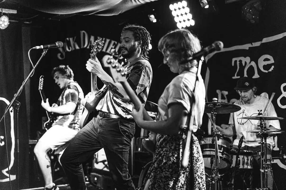 A rock band performs on stage with guitarist, bassist, vocalist and drummer under bright stage lights in a small venue.