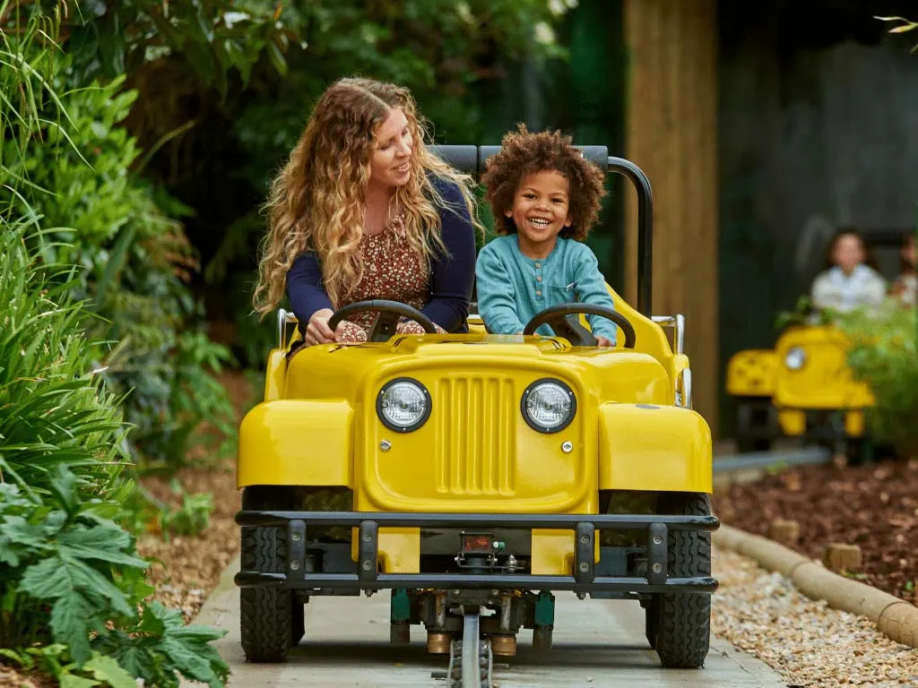 An adult and child ride together in a bright yellow miniature jeep on a track through a leafy garden setting.