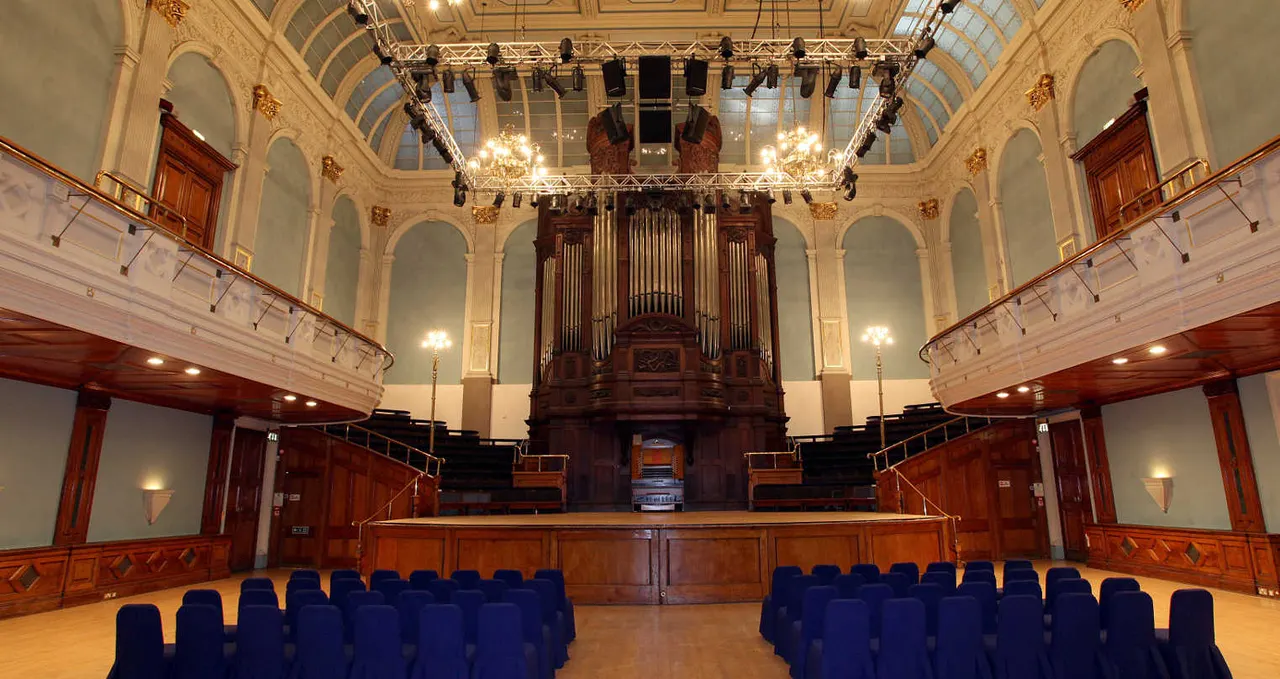 Grand ornate concert hall with blue seating facing a wooden stage and large pipe organ.