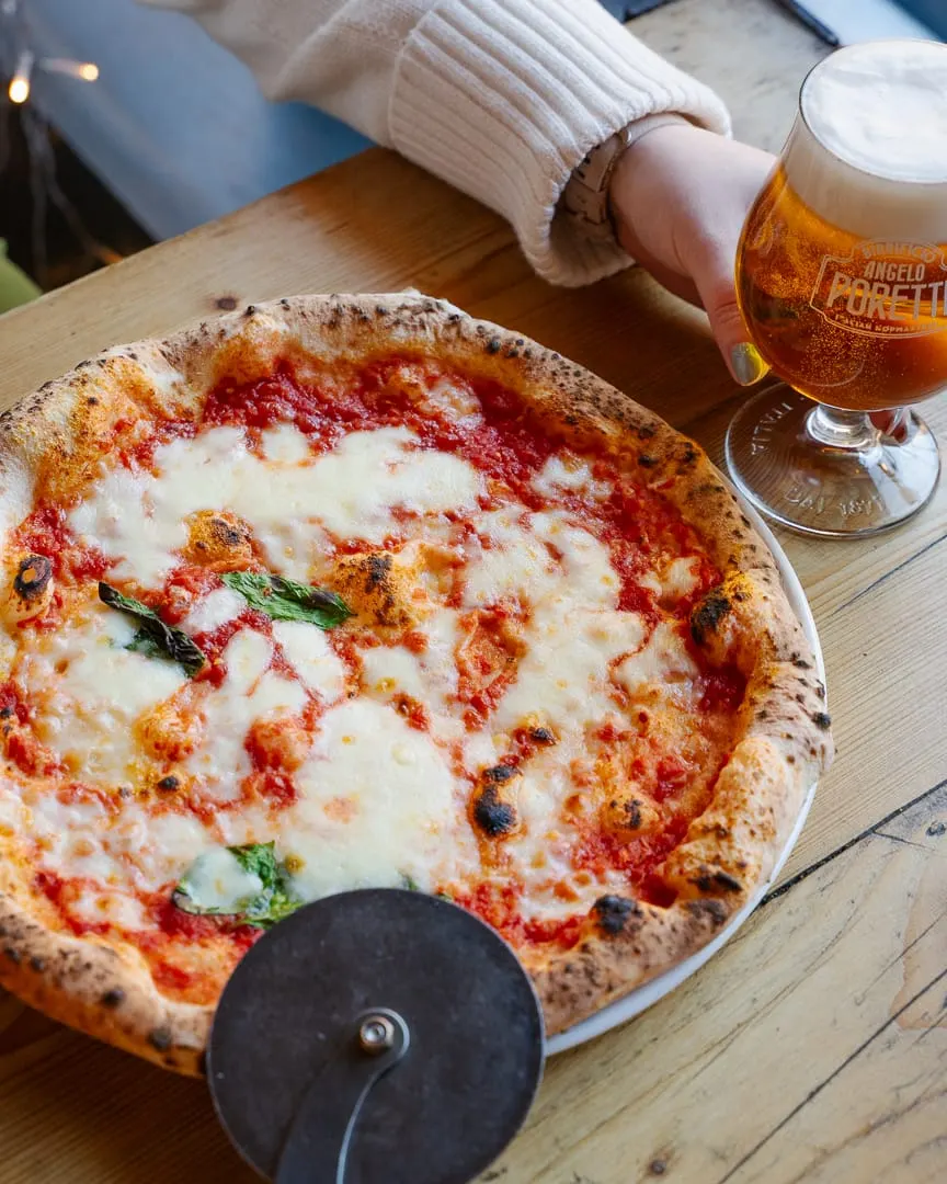 Person in cream sweater enjoying wood-fired margherita pizza with beer at a table.