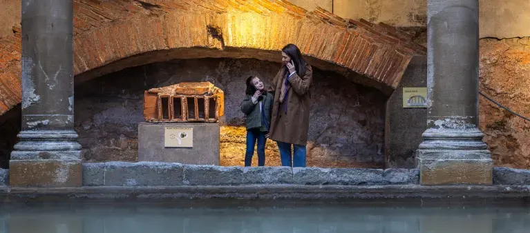 Two visitors stand beneath a brick archway beside water at an ancient Roman site, looking up at historic stonework.