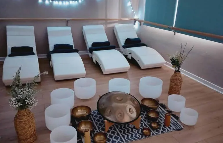 Spa relaxation room with four white loungers, white cylindrical seating, singing bowls, dried flowers, and ambient lighting.