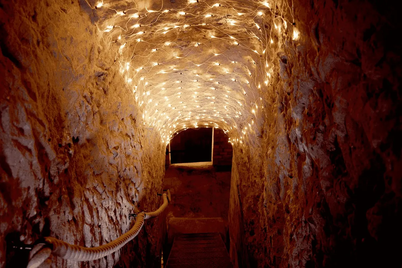 Stone tunnel lit by warm string lights with red walls and an opening ahead.
