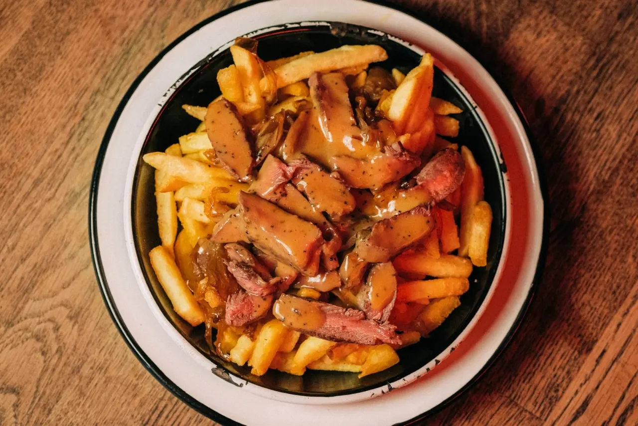 Sliced meat with caramelised onions and chips served in a black-rimmed white bowl on a wooden table.