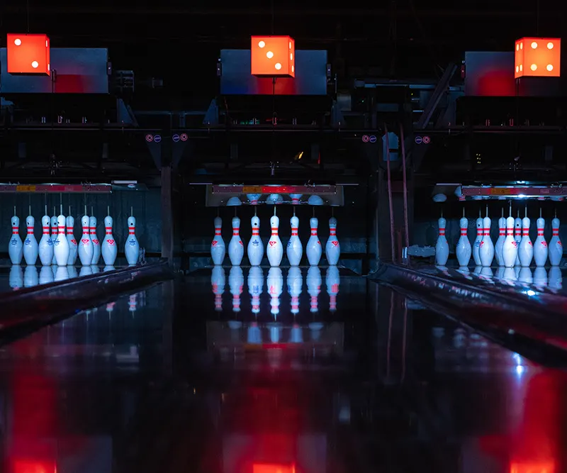 Three bowling lanes with illuminated pins and neon signage overhead at night.