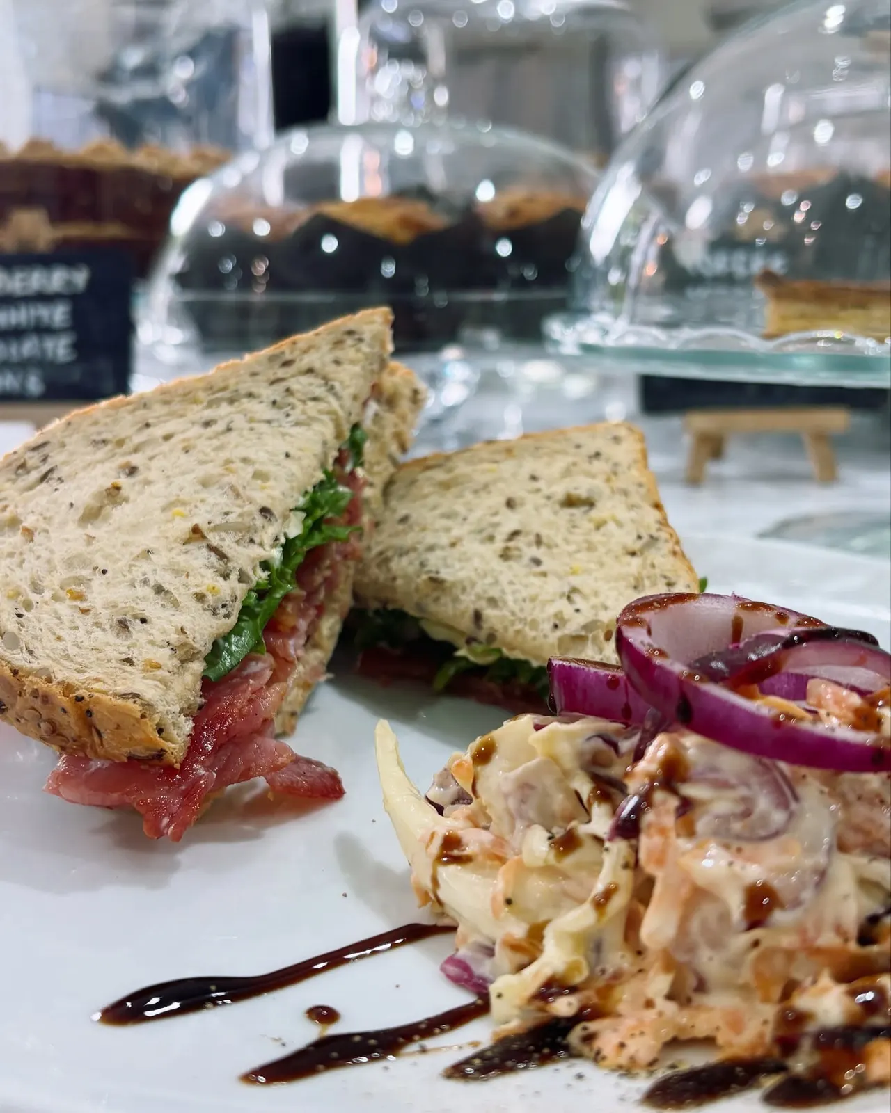 Seeded bread sandwich with bacon and leafy greens alongside a creamy spread with red onion on a white plate.