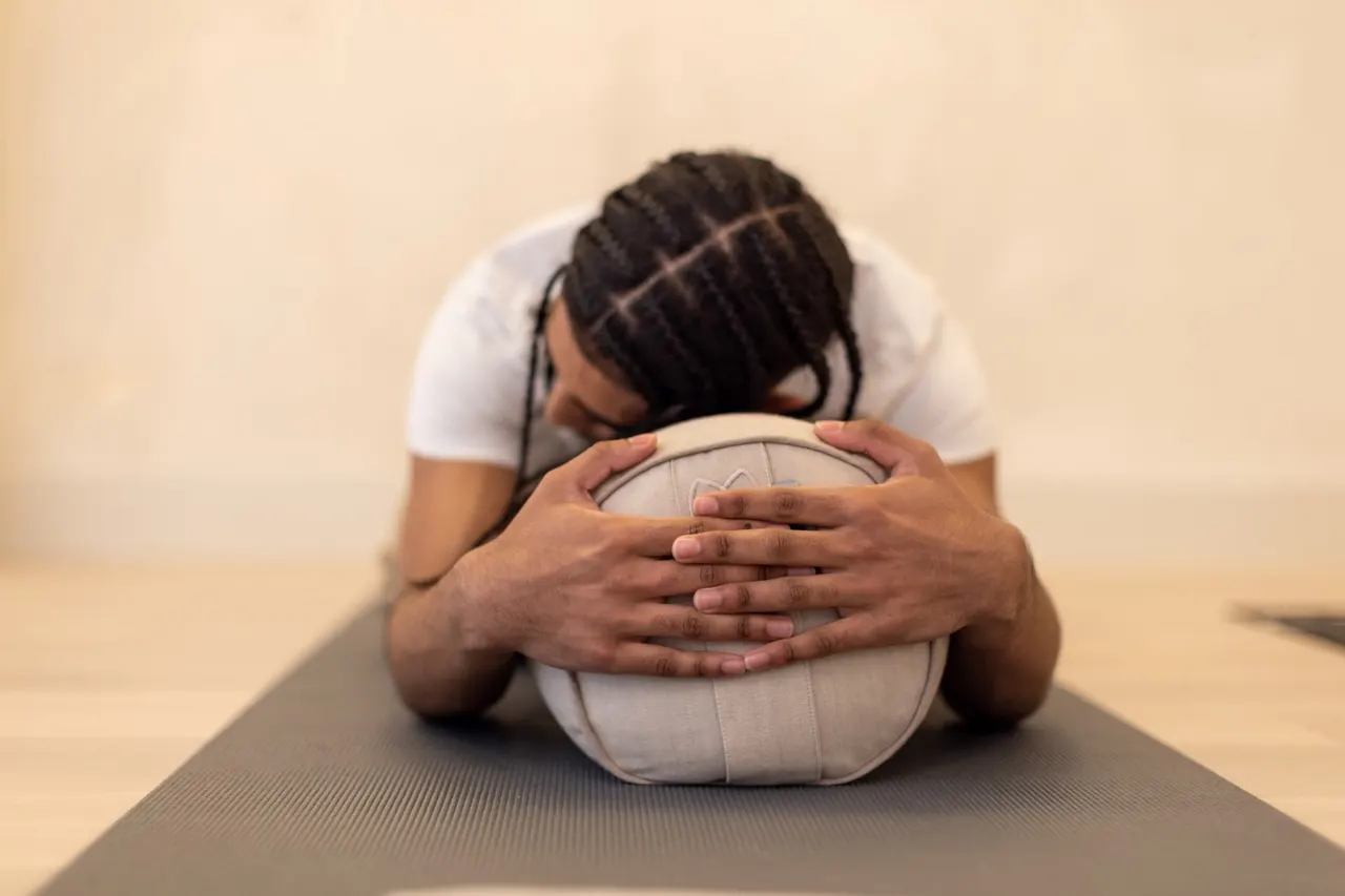 Person in white top practising forward fold yoga pose with hands clasped around volleyball on exercise mat.